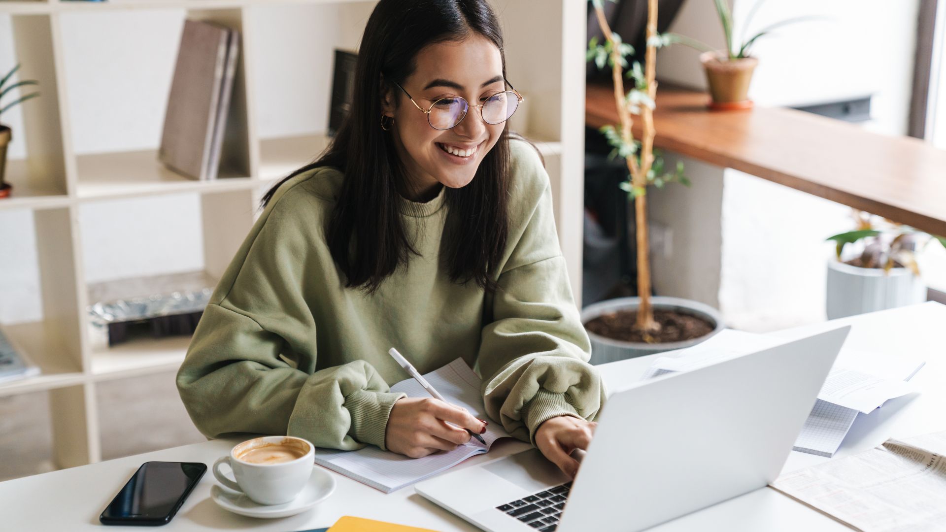 A woman wearing glasses and a green sweater is smiling while using a laptop and taking notes. A cup of coffee is on the table next to her. She's sitting in a well-lit room with plants and shelves.